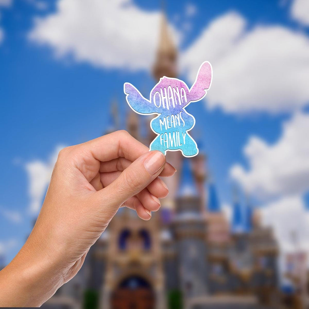 Disneyy Stitch Sticker Waterproof Disney Laptop Stickers/ Lilo and Stitch Ohana Aloha planner stationery decal water bottle cell phonelogo A Stitch-shaped sticker held in front of a blurred background of a castle and sky. The sticker is blue and pink, with the words 'Ohana Means Family' written on it in white.