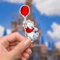 Disneyy  Winnie The Pooh Disney Laptop Sticker Positive Affirmation planner stationery decal water bottle cell.  : A sticker featuring Winnie the Pooh holding a red balloon. The bear is white with a red shirt and black outline. The balloon is a solid red circle. The background is blurred, showing a castle and blue sky with clouds. The sticker is held by a hand.