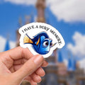A sticker featuring Dory from Finding Nemo. The blue fish has large eyes and a yellow tail. The sticker has the text 'I have a Dory memory' in a curved white banner. The background is a blurred image of a castle and blue sky.